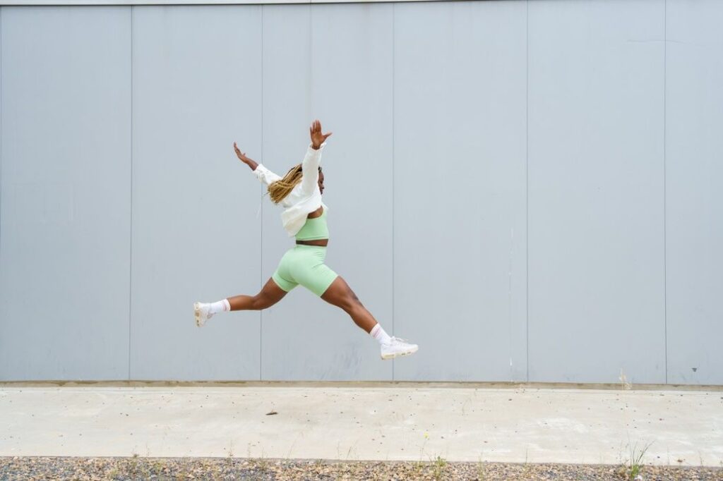 Frau springt kraftvoll in die Luft und hat grüne Sportbekleidung mit weisser Jacke und weissen Turnschuhen an.
