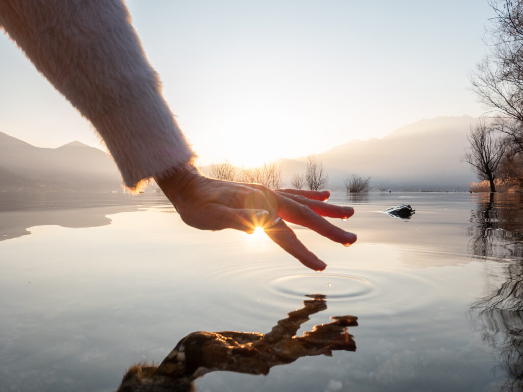 Landschaft mit Sonnenaufgang und See, wo eine Hand mit dem Zeigefinger in den See tippt.